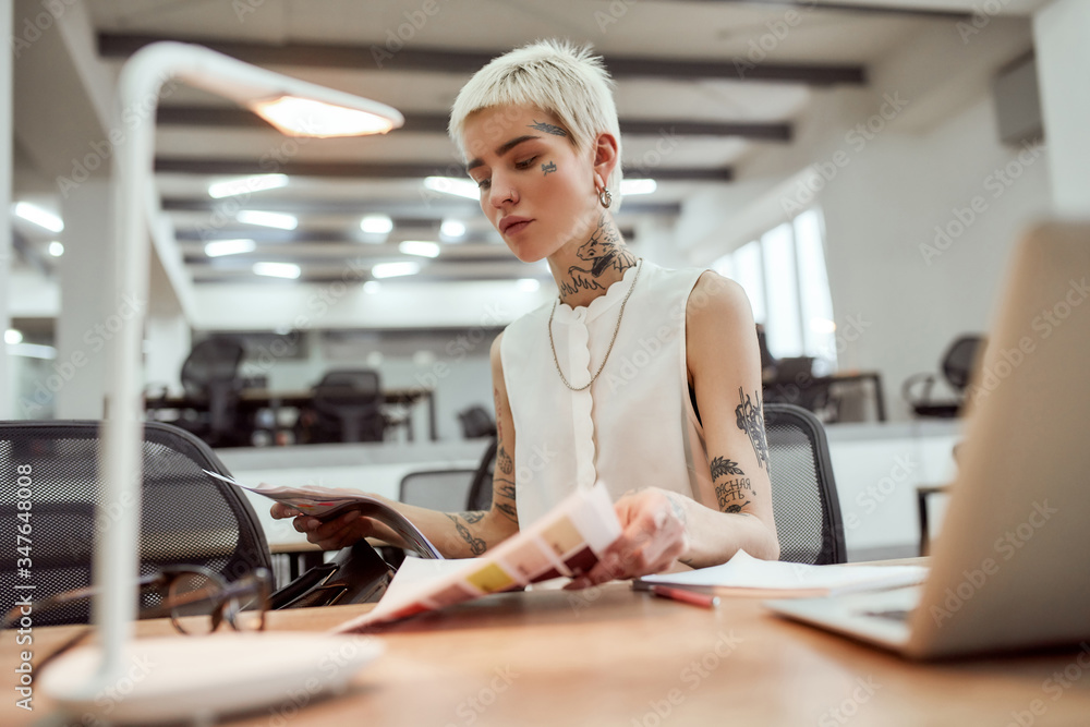 Looking for documents. Young stylish blonde tattooed businesswoman with short haircut taking out some documents from her handbag while sitting at her desk in office Looking for documents. Young stylish blonde tattooed businesswoman with short haircut taking out some documents from her handbag while sitting at her desk in office