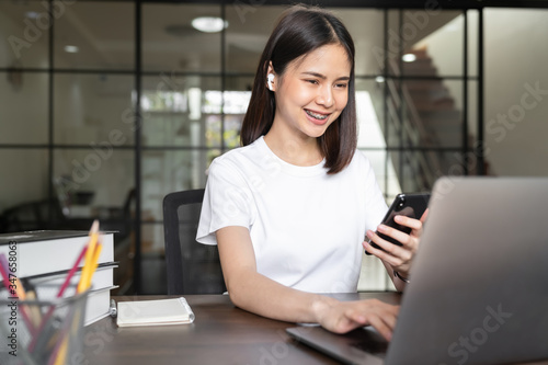 Smiley Asian woman hand holding smartphone and press the button on the keyboard laptop in house. © sompong