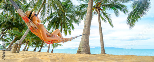 Young happy woman in hat and sunglasses laying in hammock on the tropical beach. Banner edition.