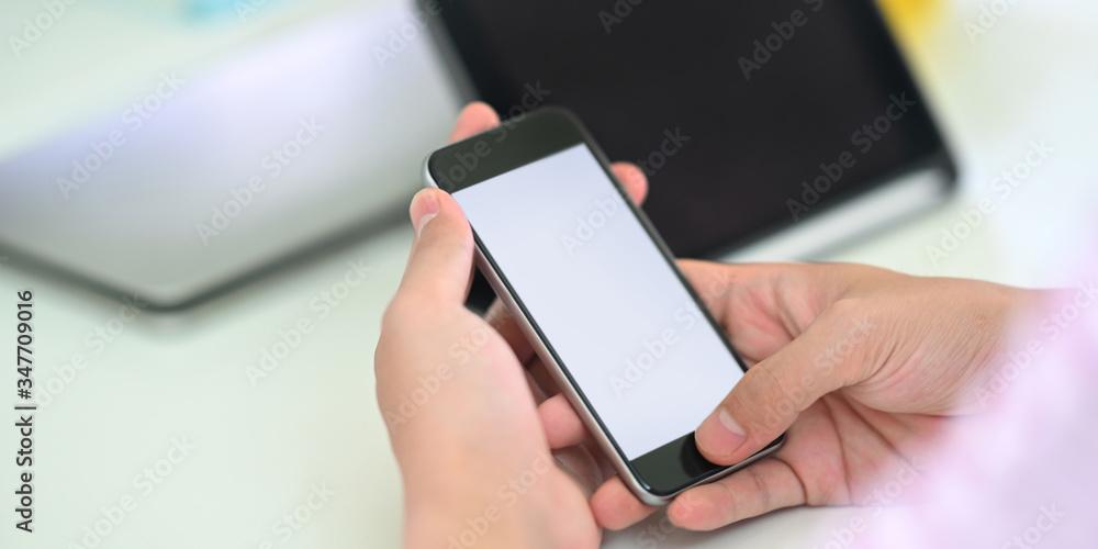 Cropped image of smart man's hands holding a white blank screen smartphone while sitting in front of computer tablet that putting on white working desk that surrounded by office equipment. Cropped image of smart man's hands holding a white blank screen smartphone while sitting in front of computer tablet that putting on white working desk that surrounded by office equipment.