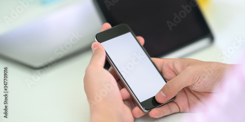 Cropped image of smart man's hands holding a white blank screen smartphone while sitting in front of computer tablet that putting on white working desk that surrounded by office equipment.