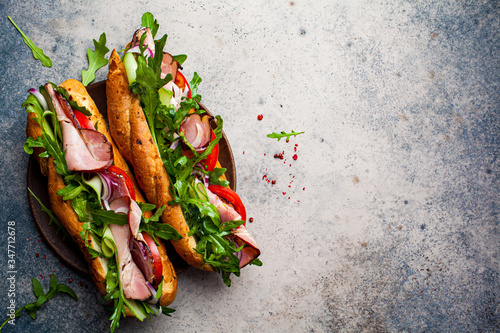 Two fresh baguette sandwiches with meat, tomato, cucumber and arugula on gray background, top view. photo