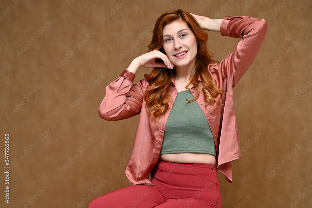 Fototapeta premium Nice girl with long hair dressed in red trousers and a pink blouse. Portrait of a young Caucasian red-haired woman sitting on a stand on a beige background in various poses.