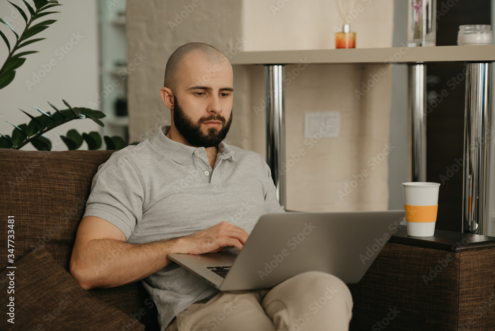 Fototapeta premium Remote work. A man working remotely on his laptop. A bald businessman with a beard works sitting on a sofa at home.