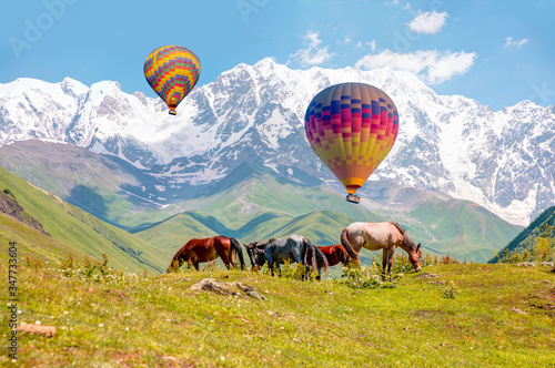 Horses grazing is green pasture against highest georgian mountain Shkhara - Colorful hot air balloon flying over Ushguli village - Upper Svaneti, Georgia