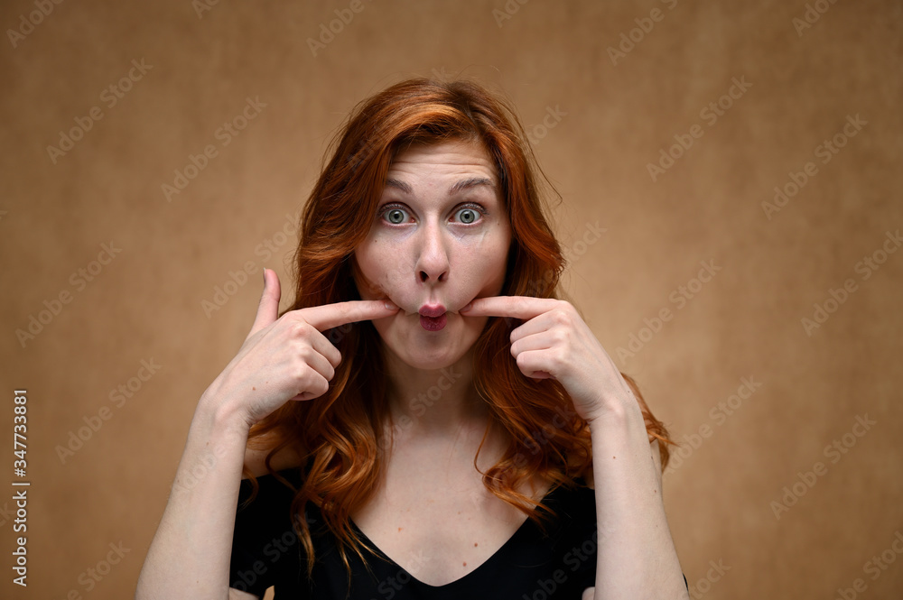 Fototapeta premium Studio large photo portrait of Caucasian young actress woman with long red hair on a beige background. Model poses with a smile and grimace.