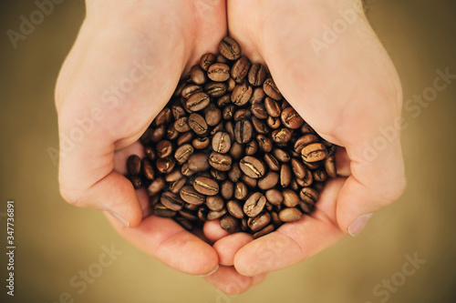 A man is holding a large handful of roasted aromatic coffee beans on a beige background, illuminated by light.