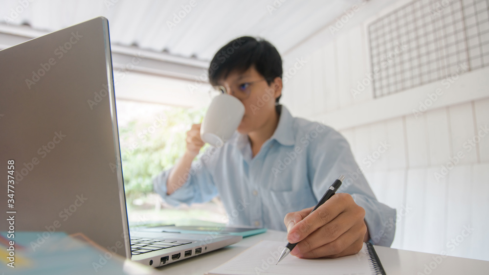 Asian young businessman of a student holding a pen writing letter on notebook and drinking hot coffee, he reading news globally on the digital laptop in the morning at home. Asian young businessman of a student holding a pen writing letter on notebook and drinking hot coffee, he reading news globally on the digital laptop in the morning at home.