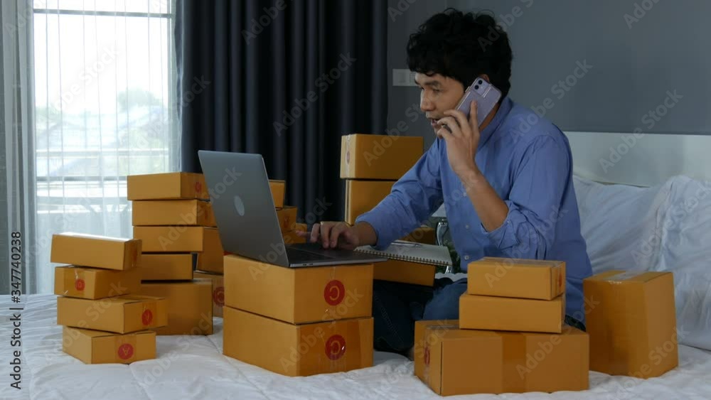 young man talking on smartphone and using laptop computer to selling product online on a bed at home young man talking on smartphone and using laptop computer to selling product online on a bed at home