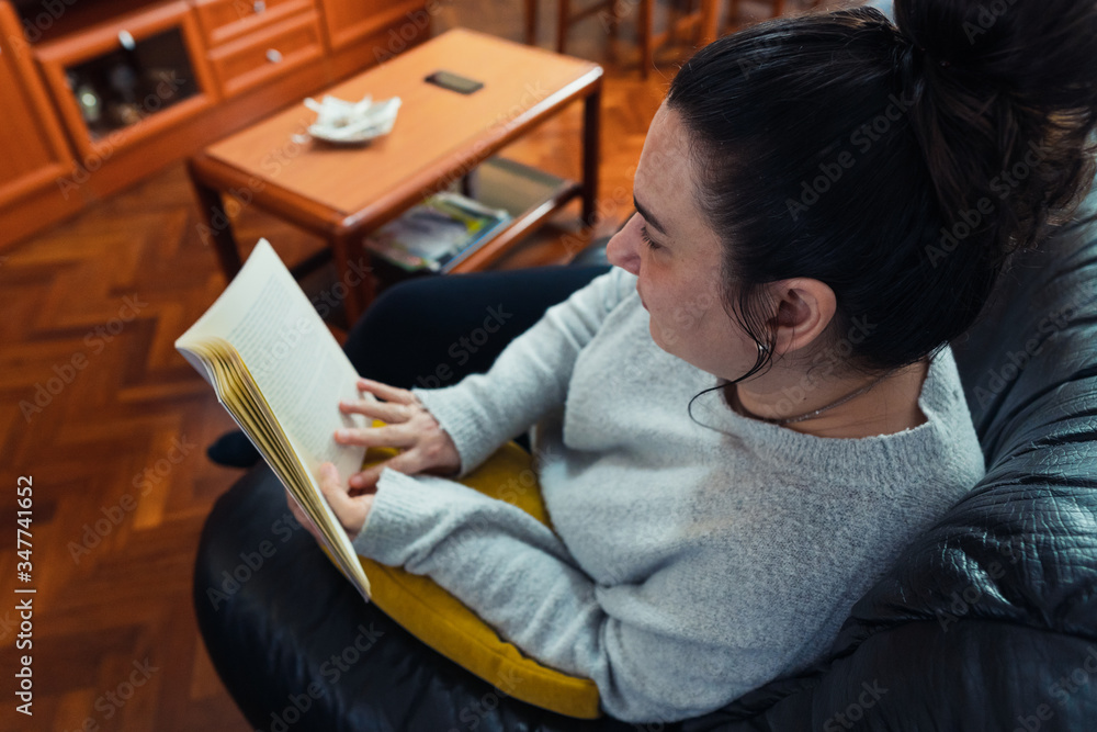 Obraz premium Middle-aged white woman reading a book lying on a black sofa with a yellow cushion in her spacious living room. She is wearing a gray sweater.