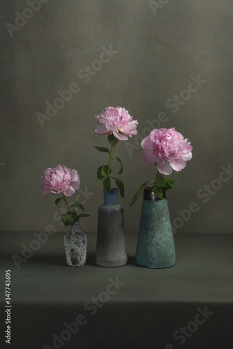 Three vintage pottery vases with pink peonies on a table in a grey room.