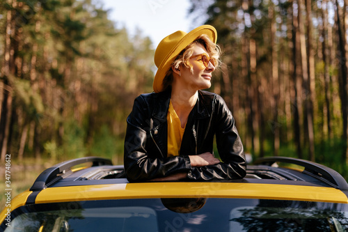 Happy Girl Enjoy Summer Sunlight at Yellow Car Sunroof, Road Trip Joy. Dreamy Woman in Jacket, Hat and Sunglasses Leaning on Auto Moon Roof at Forest. Trendy Blonde Weekend Adventure Lifestyle