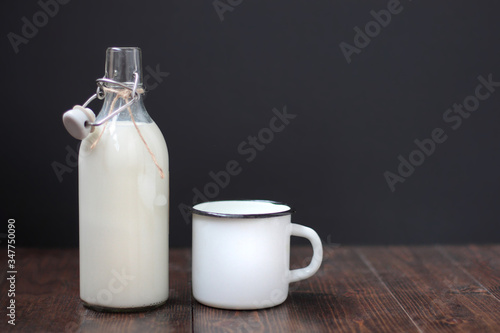 Glass bottle of milk and white metal mug on a dark rustic wooden table. Black background