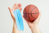 Closeup of basketball ball with a medical mask in man's hands on a light background. Cancellation of sporting events.