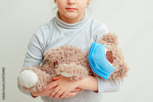 Toy bear with a medical mask in hands of sad little girl during quarantine on light background. Quarantine weekdays of children. © okskukuruza