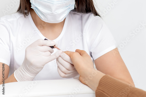 Hands of master of manicure applying nail polish on the nails of a young woman wearing protective mask in manicure salon.