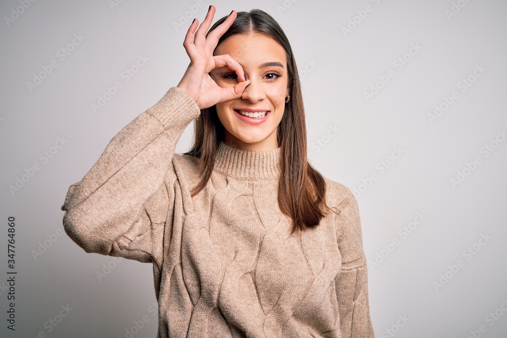 Young beautiful brunette woman wearing casual sweater standing over white background doing ok gesture with hand smiling, eye looking through fingers with happy face. Young beautiful brunette woman wearing casual sweater standing over white background doing ok gesture with hand smiling, eye looking through fingers with happy face.