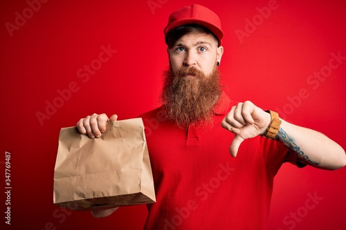 Redhead Irish delivery man with beard holding takeaway paper bag over red background with angry face, negative sign showing dislike with thumbs down, rejection concept
