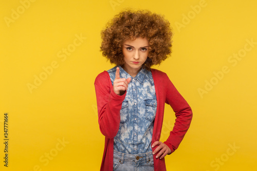 Be careful! Portrait of serious woman with curly hair in casual outfit looking displeased and giving advice, warning with admonishing finger gesture. indoor studio shot isolated on yellow background
