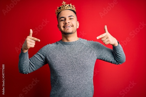 Young handsome man wearing golden crown of prince over isolated red background looking confident with smile on face, pointing oneself with fingers proud and happy.