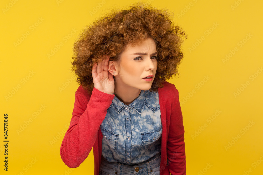 What are you saying? Portrait of woman with curly hair holding hand near ear and listening carefully intently, curious to hear secret quiet talk. indoor studio shot isolated on yellow background What are you saying? Portrait of woman with curly hair holding hand near ear and listening carefully intently, curious to hear secret quiet talk. indoor studio shot isolated on yellow background