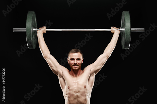 Strong smiling athletic man - athlete fitness model showing his perfect body isolated on black background with copyspace. Does an exercise, holds barbell over head, perfect abs and chest