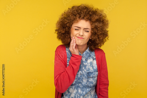 Dental problems. Portrait of unhealthy woman with curly hair wincing in pain and touching sore cheek, suffering unbearable toothache, gum disease. indoor studio shot isolated on yellow background © khosrork