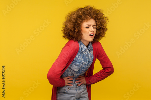 Abdominal pain. Portrait of sick woman with curly hair wincing in pain and clutching belly, suffering severe stomach ache, period spasm and cramps. indoor studio shot isolated on yellow background photo