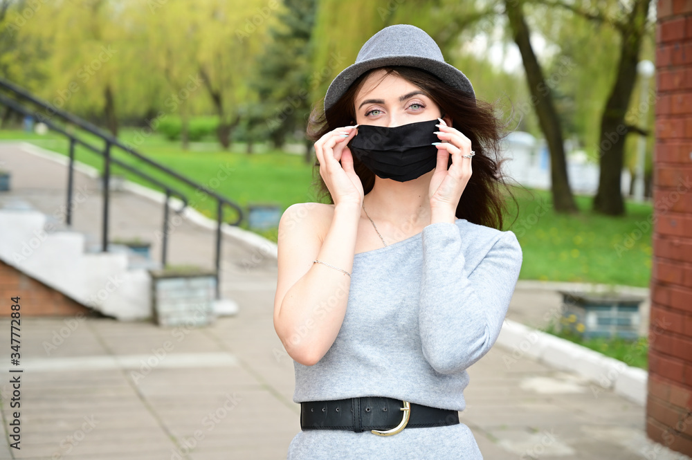 Model posing in a city park outdoors. Photo of a cute young Caucasian brunette girl in a hat in a gray dress in black medical mask.