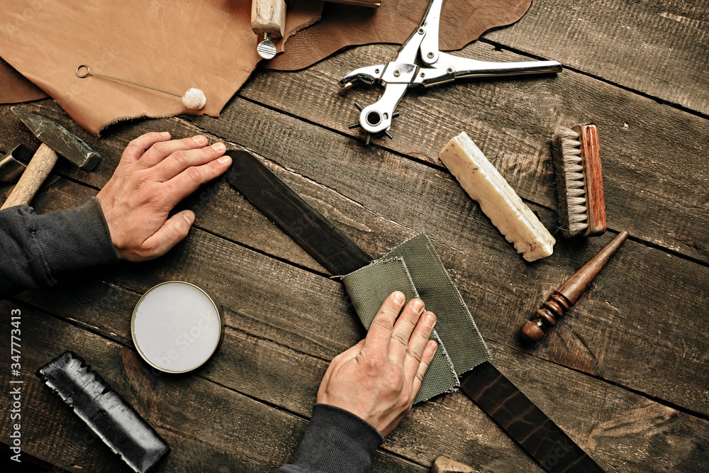 Working process of the leather belt in leather workshop. Man holding tool. Tanner in old tannery. Wooden table background. Close up man arm. Maintenance concept. Goods production. Top view, flat lay.