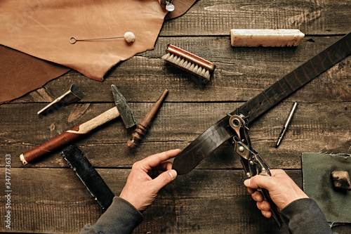 Working process of the leather belt in leather workshop. Man holding tool. Tanner in old tannery. Wooden table background. Close up man arm. Maintenance concept. Goods products. Top view, flat lay. photo