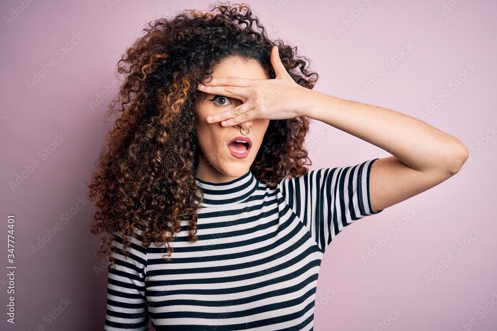 Young beautiful woman with curly hair and piercing wearing casual striped t-shirt peeking in shock covering face and eyes with hand, looking through fingers with embarrassed expression. Young beautiful woman with curly hair and piercing wearing casual striped t-shirt peeking in shock covering face and eyes with hand, looking through fingers with embarrassed expression.