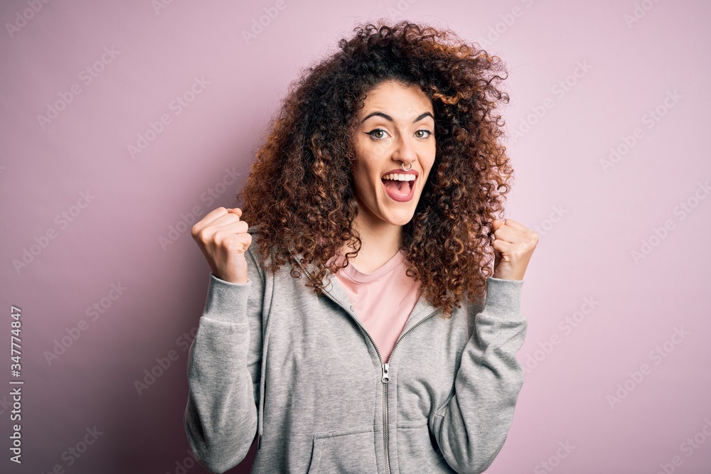 Young beautiful brunette sportswoman with curly hair and piercing wearing sportswear celebrating surprised and amazed for success with arms raised and open eyes. Winner concept. Young beautiful brunette sportswoman with curly hair and piercing wearing sportswear celebrating surprised and amazed for success with arms raised and open eyes. Winner concept.