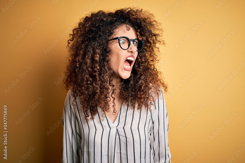 Young beautiful woman with curly hair and piercing wearing striped shirt and glasses angry and mad screaming frustrated and furious, shouting with anger. Rage and aggressive concept. Young beautiful woman with curly hair and piercing wearing striped shirt and glasses angry and mad screaming frustrated and furious, shouting with anger. Rage and aggressive concept.