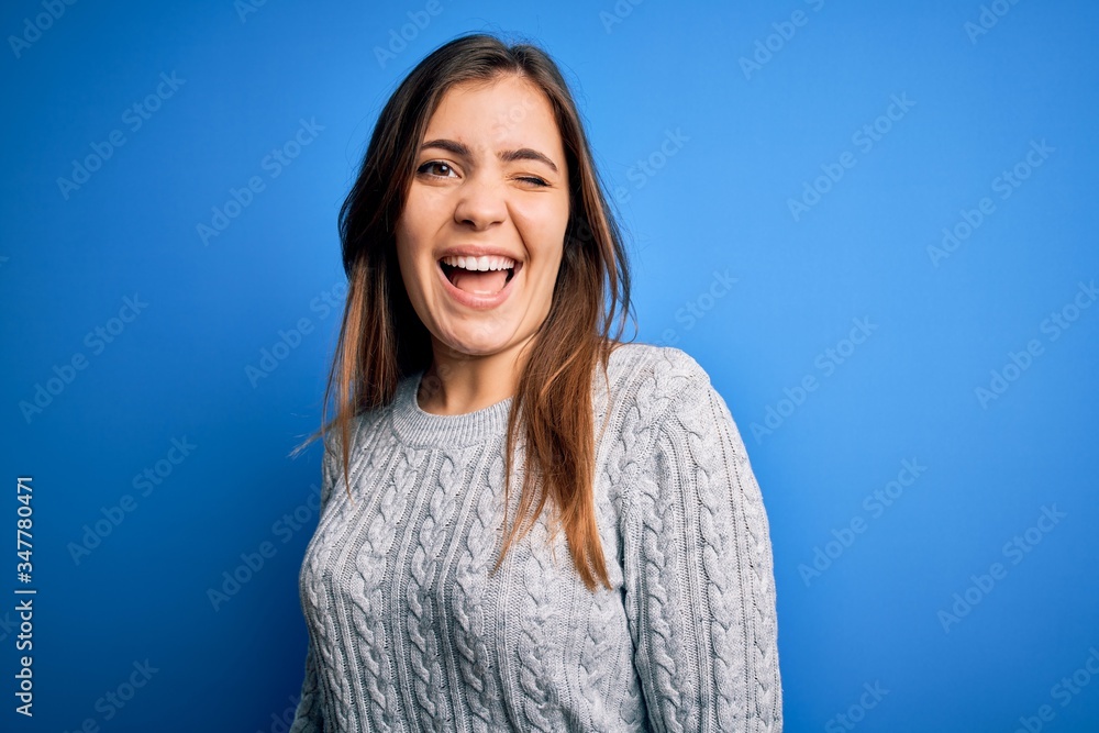 Beautiful young woman wearing casual wool sweater standing over blue isolated background winking looking at the camera with sexy expression, cheerful and happy face. Beautiful young woman wearing casual wool sweater standing over blue isolated background winking looking at the camera with sexy expression, cheerful and happy face.