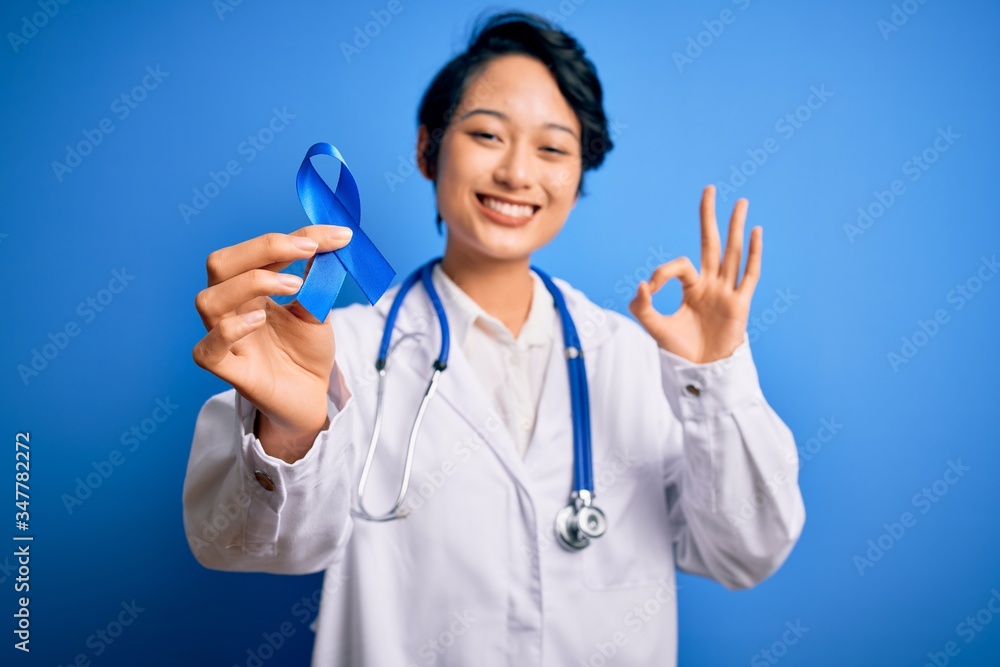Young beautiful asian doctor girl wearing stethoscope and coat holding blue cancer ribbon doing ok sign with fingers, excellent symbol Young beautiful asian doctor girl wearing stethoscope and coat holding blue cancer ribbon doing ok sign with fingers, excellent symbol