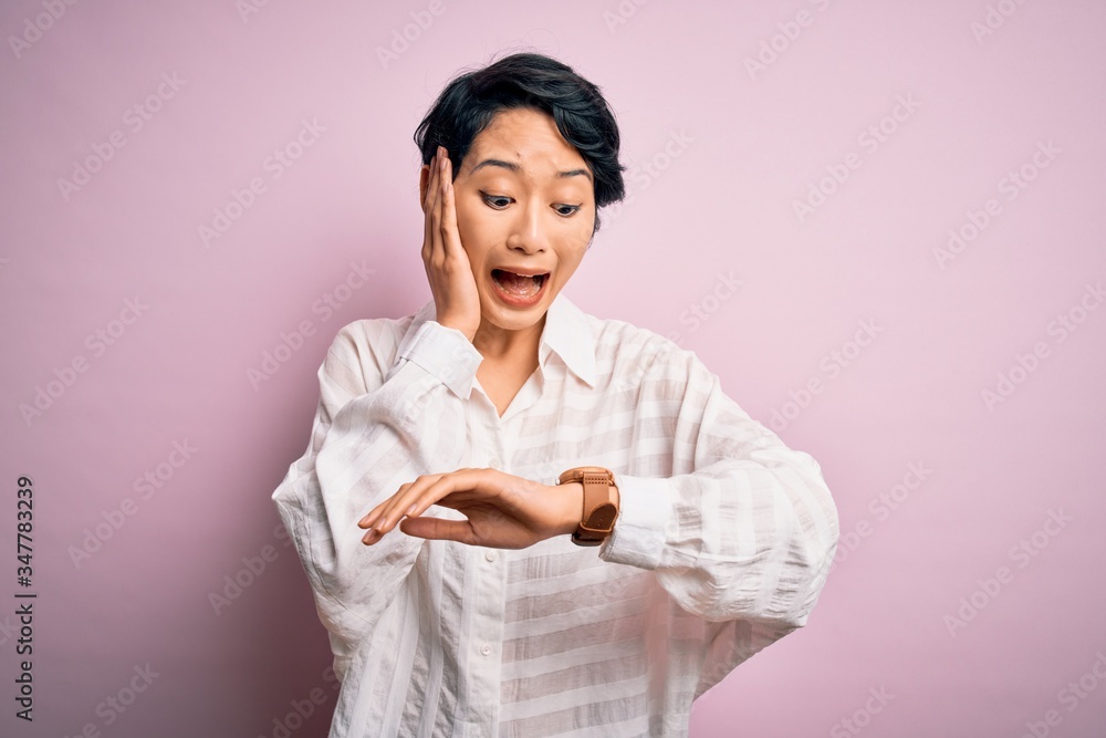 Young beautiful asian girl wearing casual shirt standing over isolated pink background Looking at the watch time worried, afraid of getting late