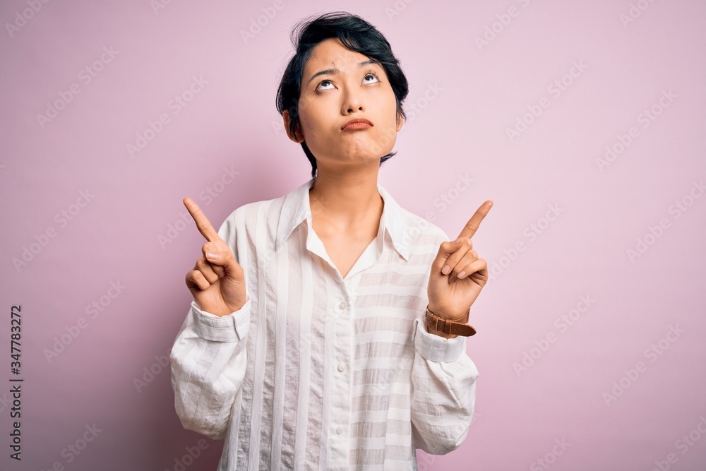 Young beautiful asian girl wearing casual shirt standing over isolated pink background Pointing up looking sad and upset, indicating direction with fingers, unhappy and depressed. Young beautiful asian girl wearing casual shirt standing over isolated pink background Pointing up looking sad and upset, indicating direction with fingers, unhappy and depressed.