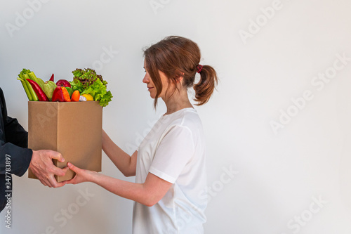 Food delivery service. A courier hands a cardboard box of fresh and raw vegetables and fruits to a young woman on a white background. Copy space.