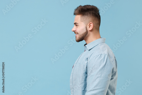 Side view of smiling young bearded guy 20s in casual shirt posing isolated on pastel blue wall background studio portrait. People sincere emotions lifestyle concept. Mock up copy space. Looking aside.