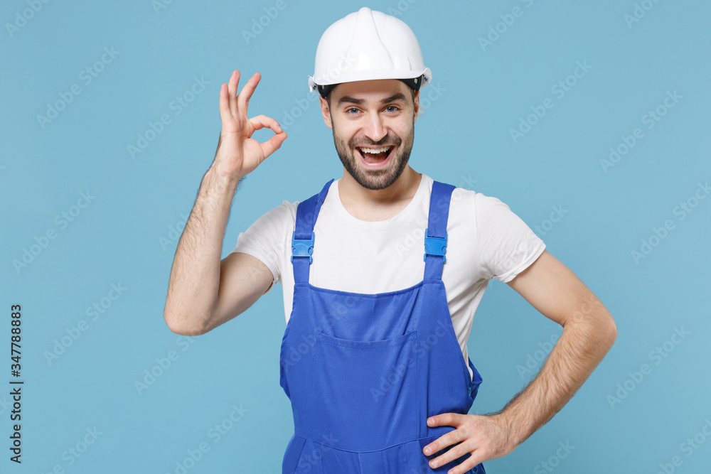 Cheerful young man in coveralls protective helmet hardhat isolated on blue background studio portrait. Instruments accessories for renovation apartment room. Repair home concept. Showing Ok gesture.