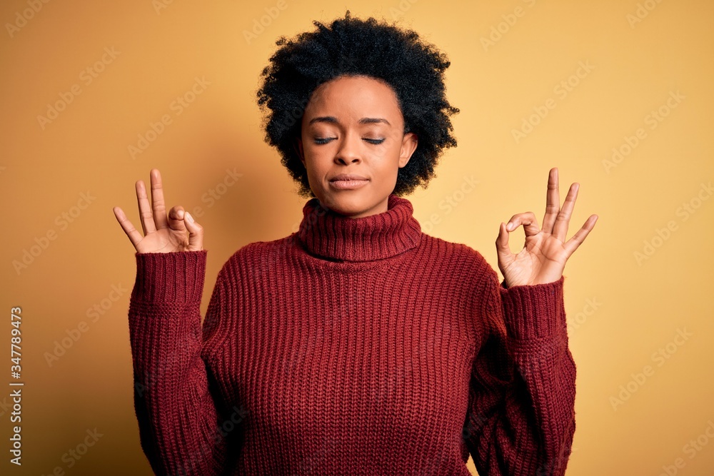 Young beautiful African American afro woman with curly hair wearing casual turtleneck sweater relaxed and smiling with eyes closed doing meditation gesture with fingers. Yoga concept. Young beautiful African American afro woman with curly hair wearing casual turtleneck sweater relaxed and smiling with eyes closed doing meditation gesture with fingers. Yoga concept.
