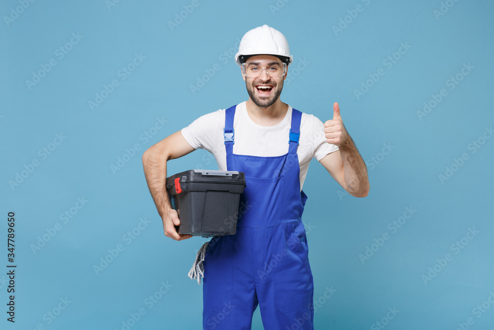 Excited man in coveralls protective helmet hardhat glasses hold tool case box isolated on blue background. Instruments accessories for renovation apartment room. Repair home concept. Showing thumb up. Excited man in coveralls protective helmet hardhat glasses hold tool case box isolated on blue background. Instruments accessories for renovation apartment room. Repair home concept. Showing thumb up.