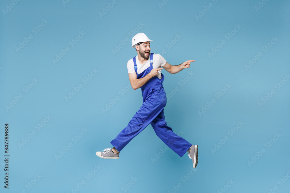 Side view of surprised man in coveralls protective helmet hardhat isolated on blue background. Instruments accessories renovation apartment room. Repair home concept. Jump point index fingers aside. Side view of surprised man in coveralls protective helmet hardhat isolated on blue background. Instruments accessories renovation apartment room. Repair home concept. Jump point index fingers aside.