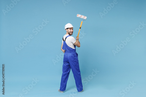 Back rear view of cheerful young man in coveralls protective helmet hardhat hold paint roller isolated on blue background. Instruments accessories for renovation apartment room. Repair home concept.