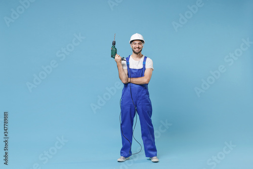Smiling young man in coveralls protective helmet hardhat glasses hold electric drill isolated on blue background in studio. Instruments accessories for renovation apartment room. Repair home concept.