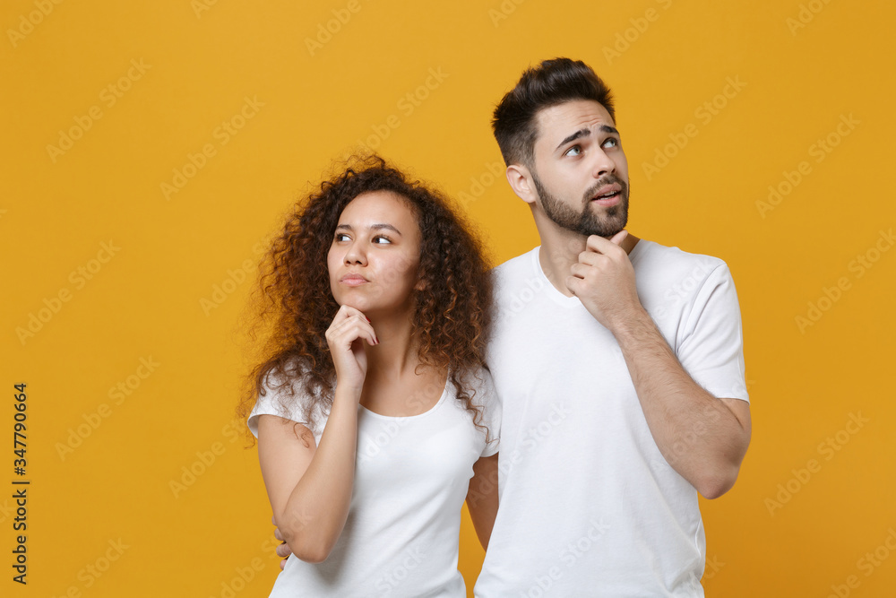 Pensive young couple two friends european guy african american girl in white t-shirts posing isolated on yellow wall background. People lifestyle concept. Put hands prop up on chin, looking aside up. Pensive young couple two friends european guy african american girl in white t-shirts posing isolated on yellow wall background. People lifestyle concept. Put hands prop up on chin, looking aside up.