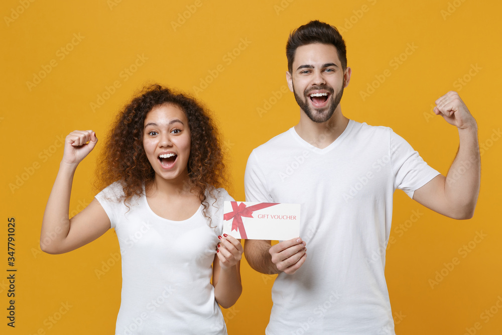 Excited young couple two friends european guy african american girl in white t-shirts posing isolated on yellow background studio. People lifestyle concept. Hold gift certificate doing winner gesture. Excited young couple two friends european guy african american girl in white t-shirts posing isolated on yellow background studio. People lifestyle concept. Hold gift certificate doing winner gesture.