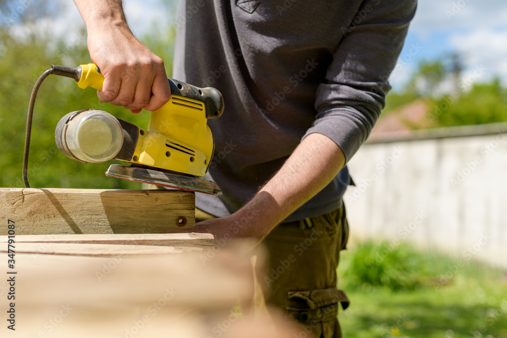 Unrecognizable man in the garden sanding wooden planks. DIY home improvement, restoration, carpentry concept. Midsection hand detail shot.