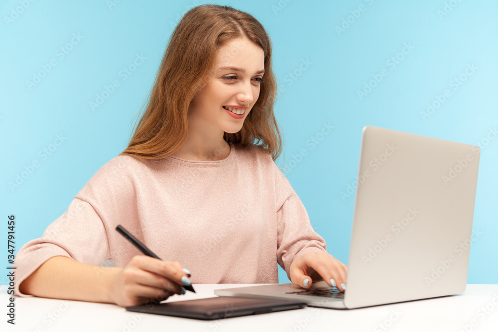 Charming smiling woman, creative designer using graphic tablet and typing keyboard laptop, working on professional digital art equipment to draw sketch. indoor studio shot isolated on blue background Charming smiling woman, creative designer using graphic tablet and typing keyboard laptop, working on professional digital art equipment to draw sketch. indoor studio shot isolated on blue background